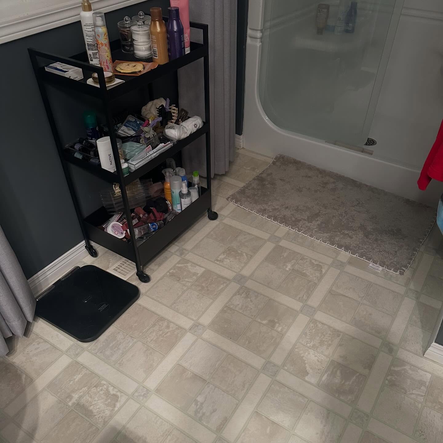 Bathroom before the floor refresh, with dated beige vinyl tile, a black rolling cart, and a white tub against deep grey walls