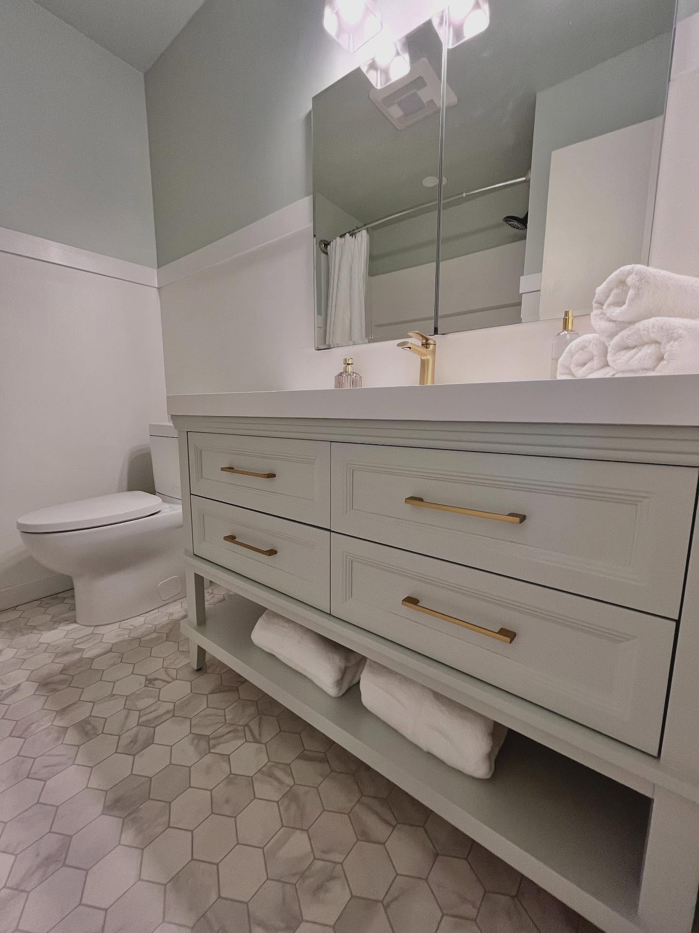 Finished bathroom vanity with a light grey four-drawer cabinet, white counter, warm brass faucet and pulls, and hexagonal marble-look floor tile