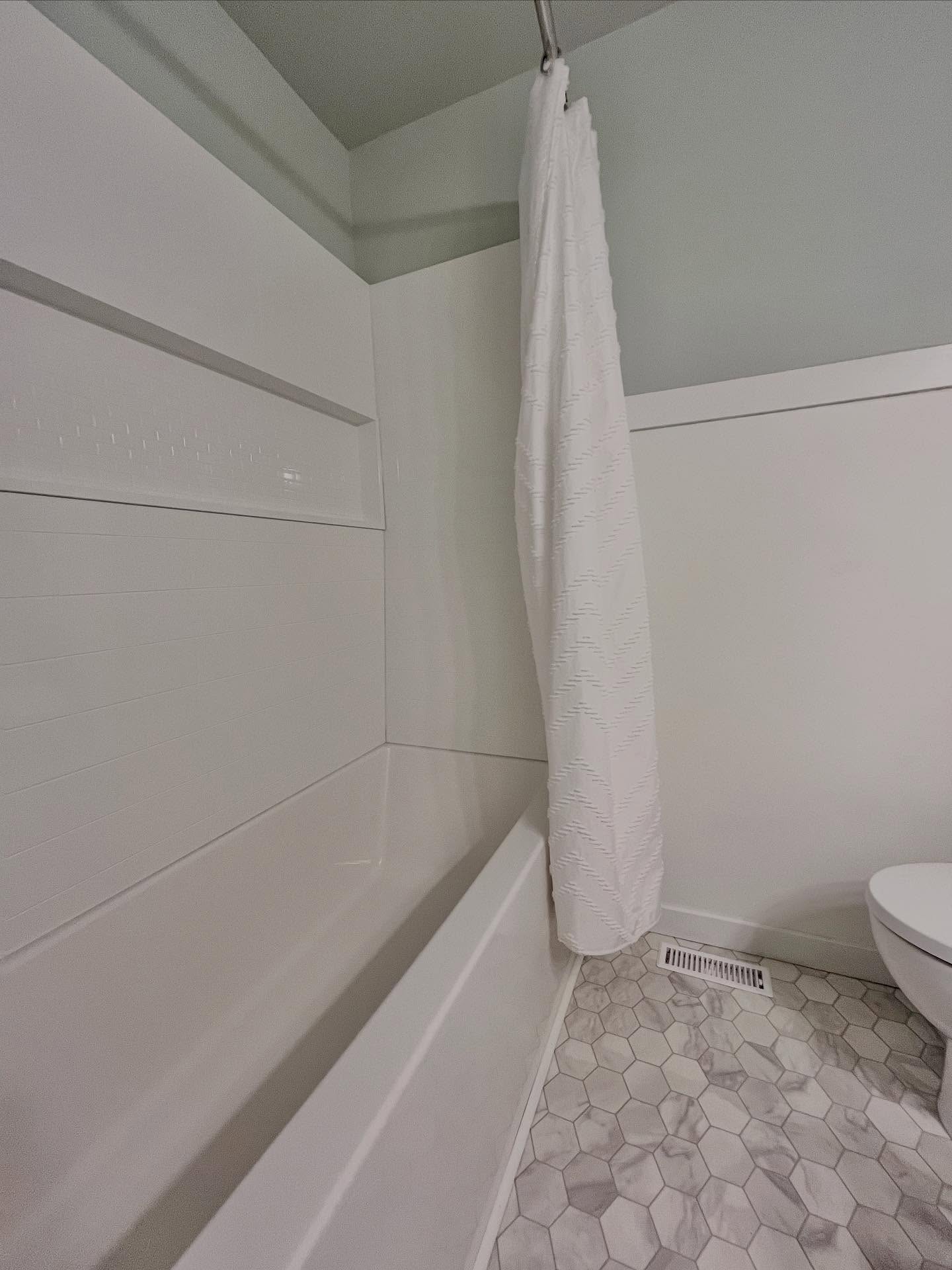 Finished white alcove tub with a recessed niche, white shower curtain, hexagonal marble-look floor, and pale sage walls