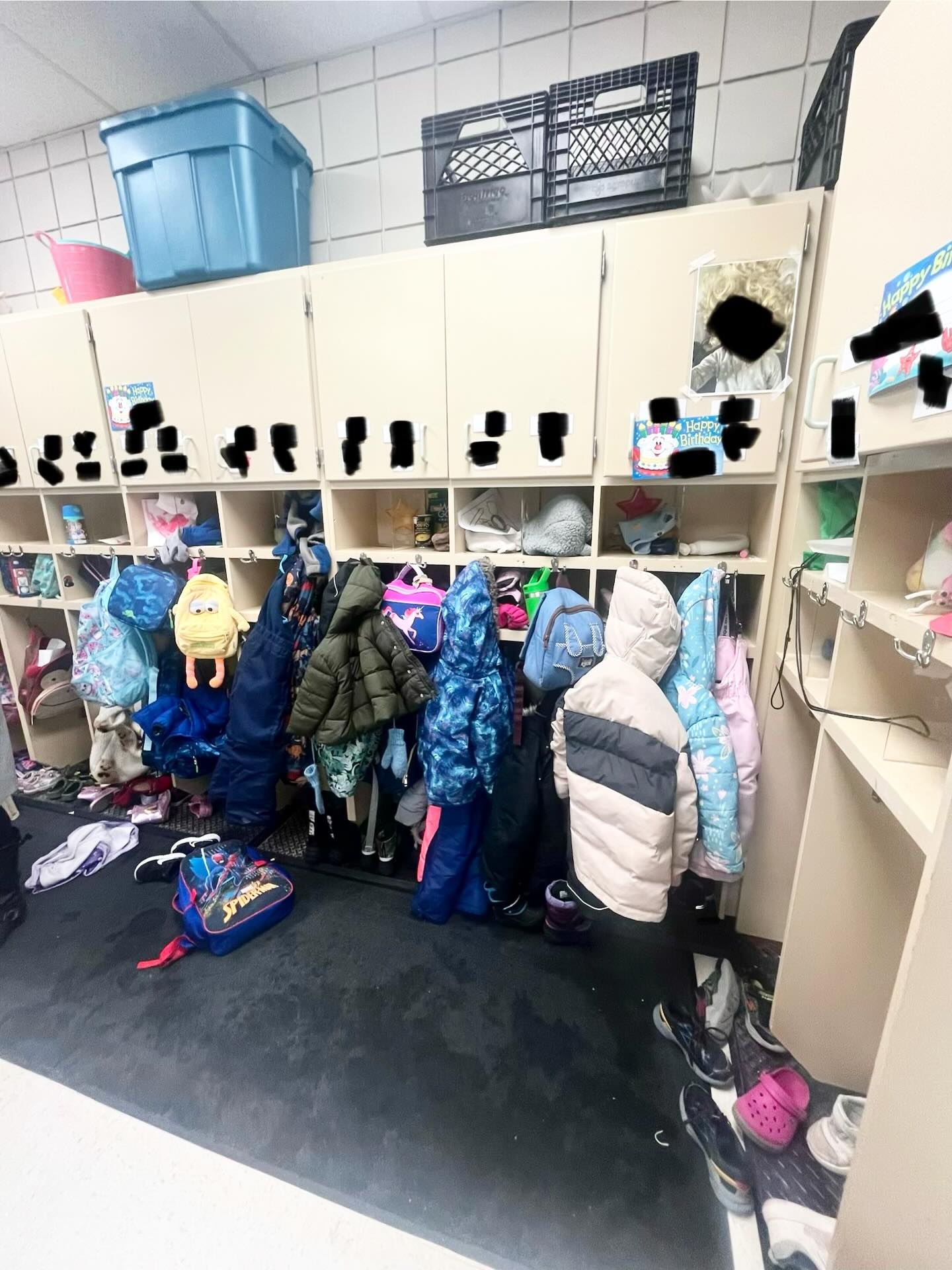 Daycare locker wall before renovation, with closed upper cabinets, cubbies, hanging stalls, coats, and bins stacked on top