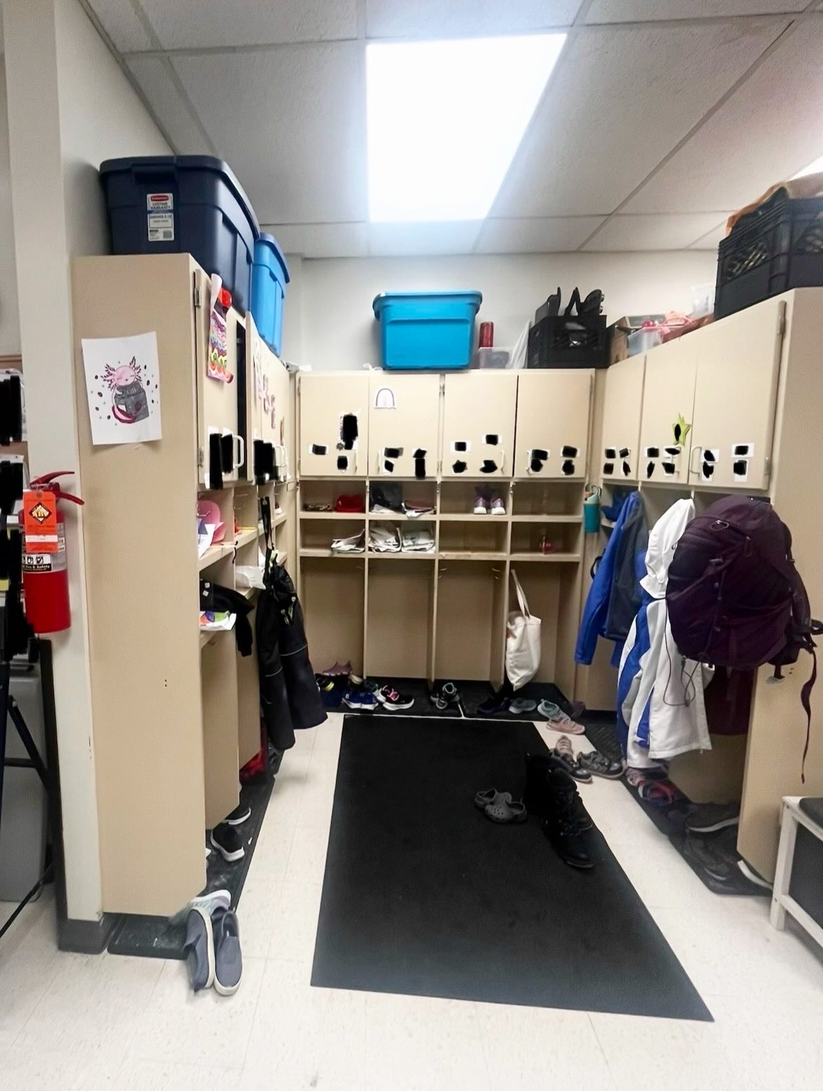 Daycare storage alcove before renovation, with beige lockers on three sides, stacked bins, and children's coats and shoes