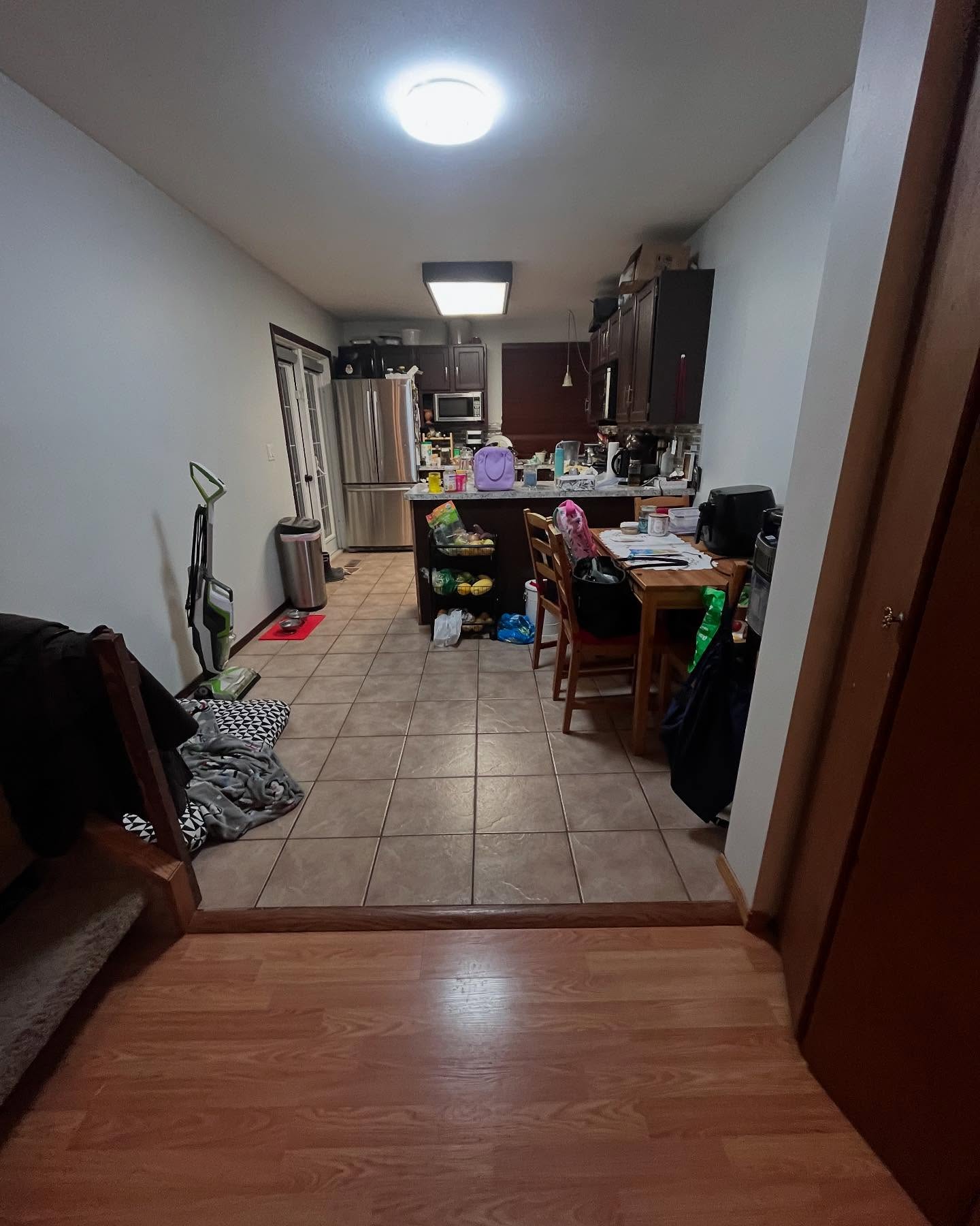 Project Daisy kitchen from the adjoining room before renovation, with dark wood cabinets, tan floor tile, and a crowded walkway