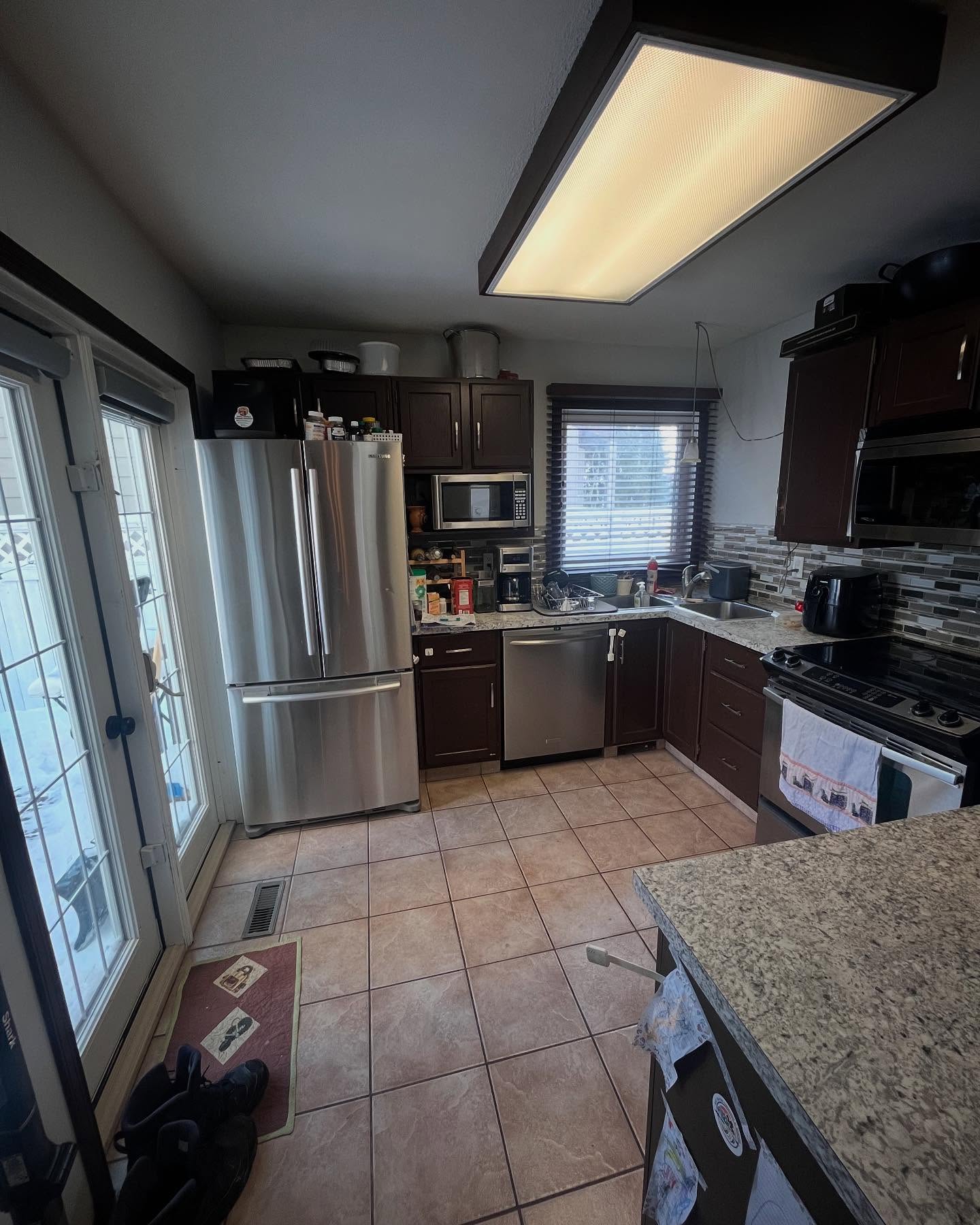 Project Daisy kitchen before renovation, with dark wood cabinets, a mosaic backsplash, stainless appliances, and tan tile floors