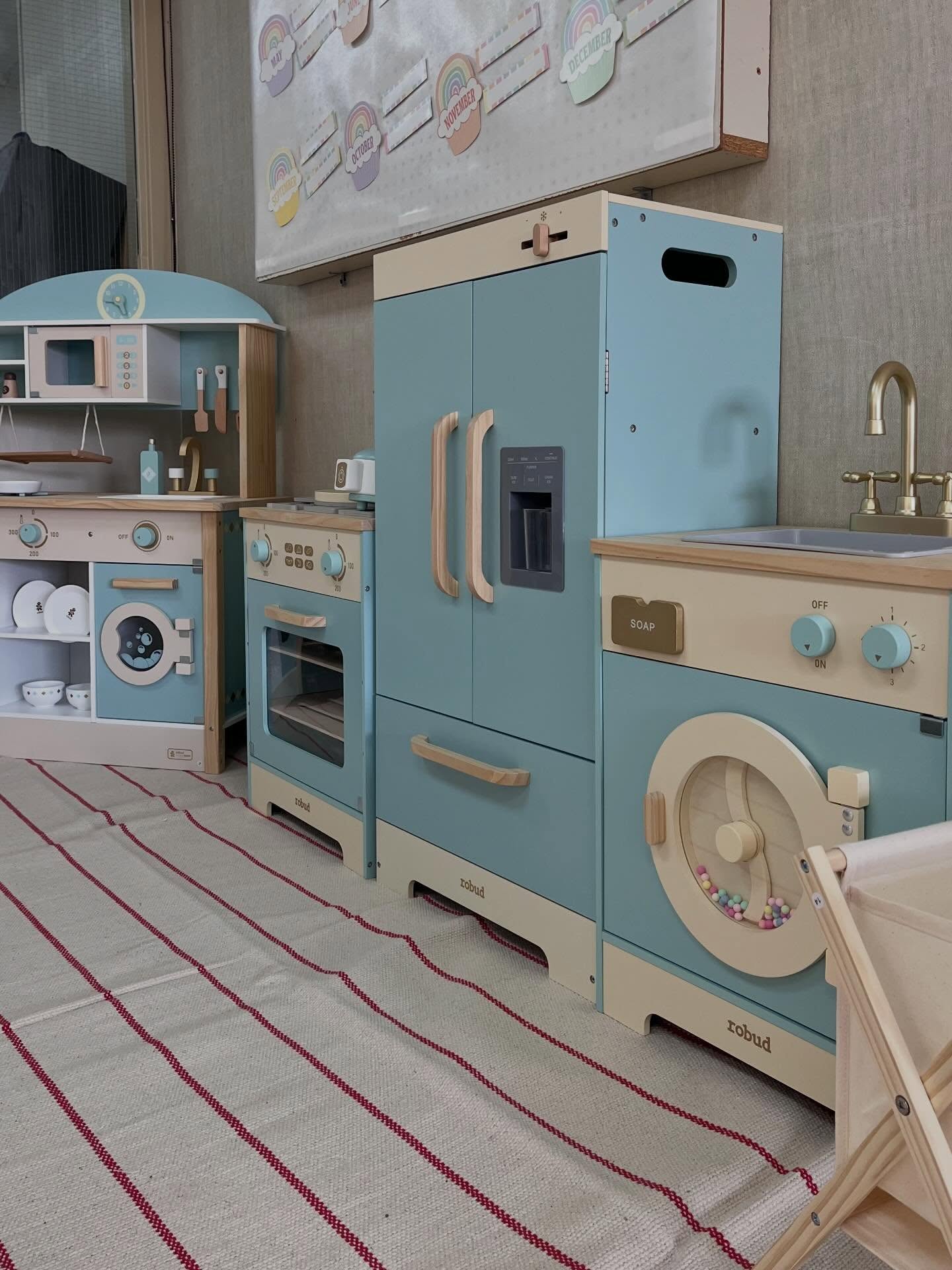 Finished wooden play kitchen in a daycare room, with oven, refrigerator, and sink units in soft blue and cream