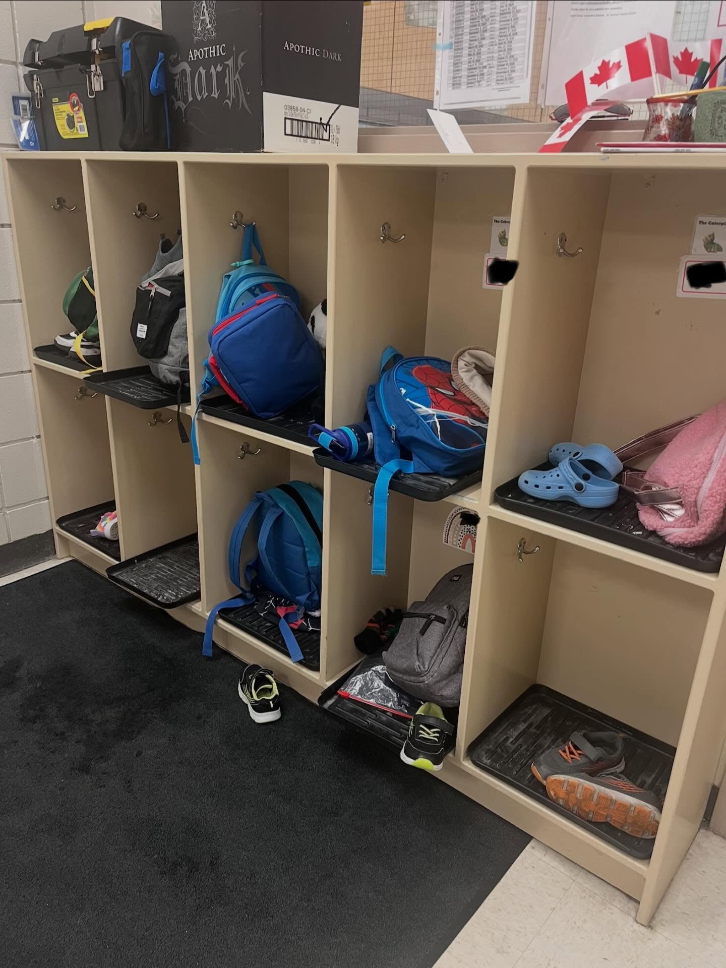 Open beige daycare locker cubbies in use, with backpacks, shoes, trays, and boxes stored on top