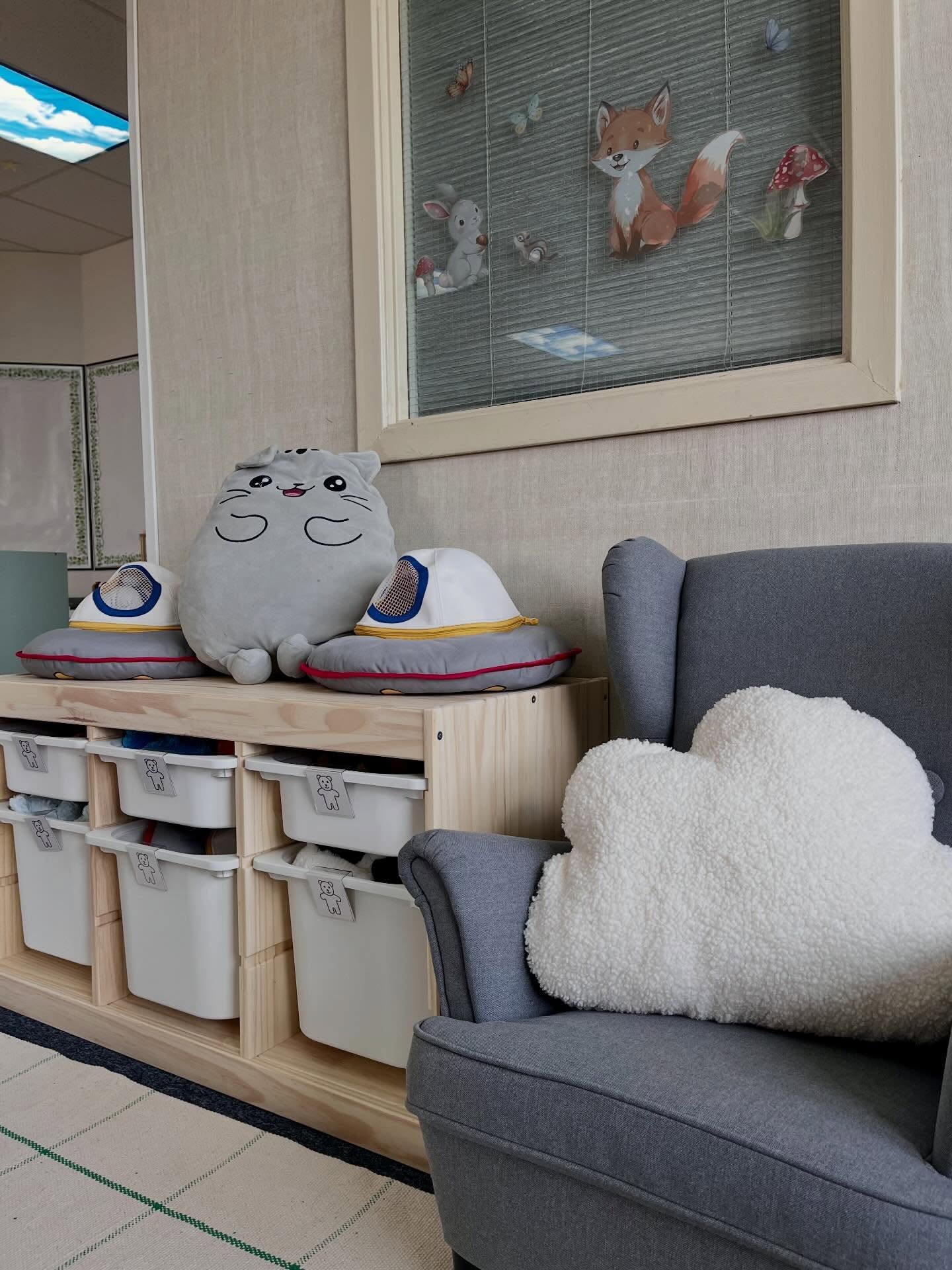 Finished daycare reading corner with labeled white bins, a grey armchair, a cloud pillow, and a window with woodland decals