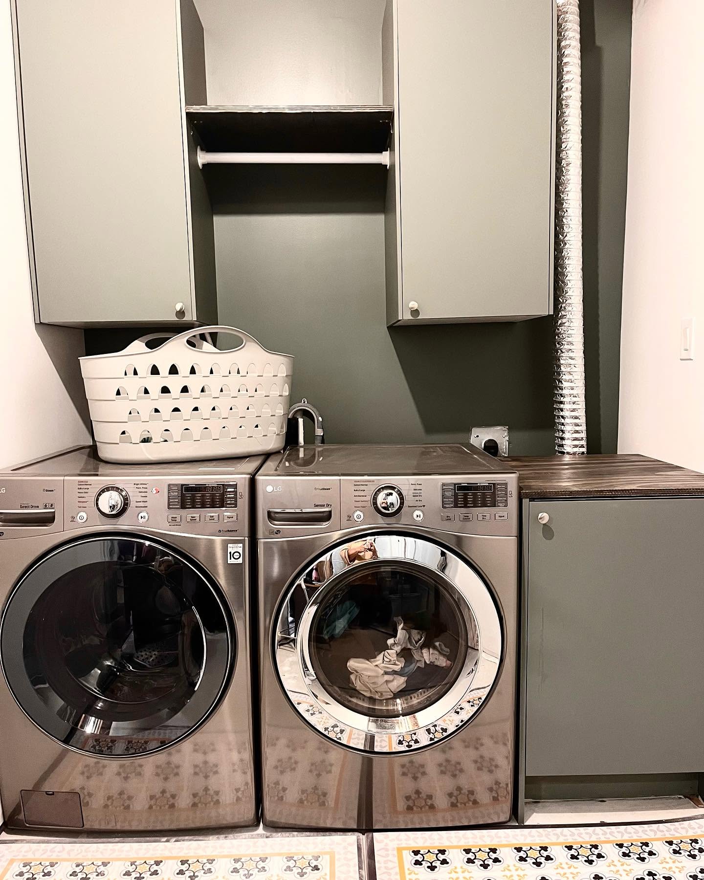 Laundry room with paired front-load machines, sage cabinets, and a forest green wall