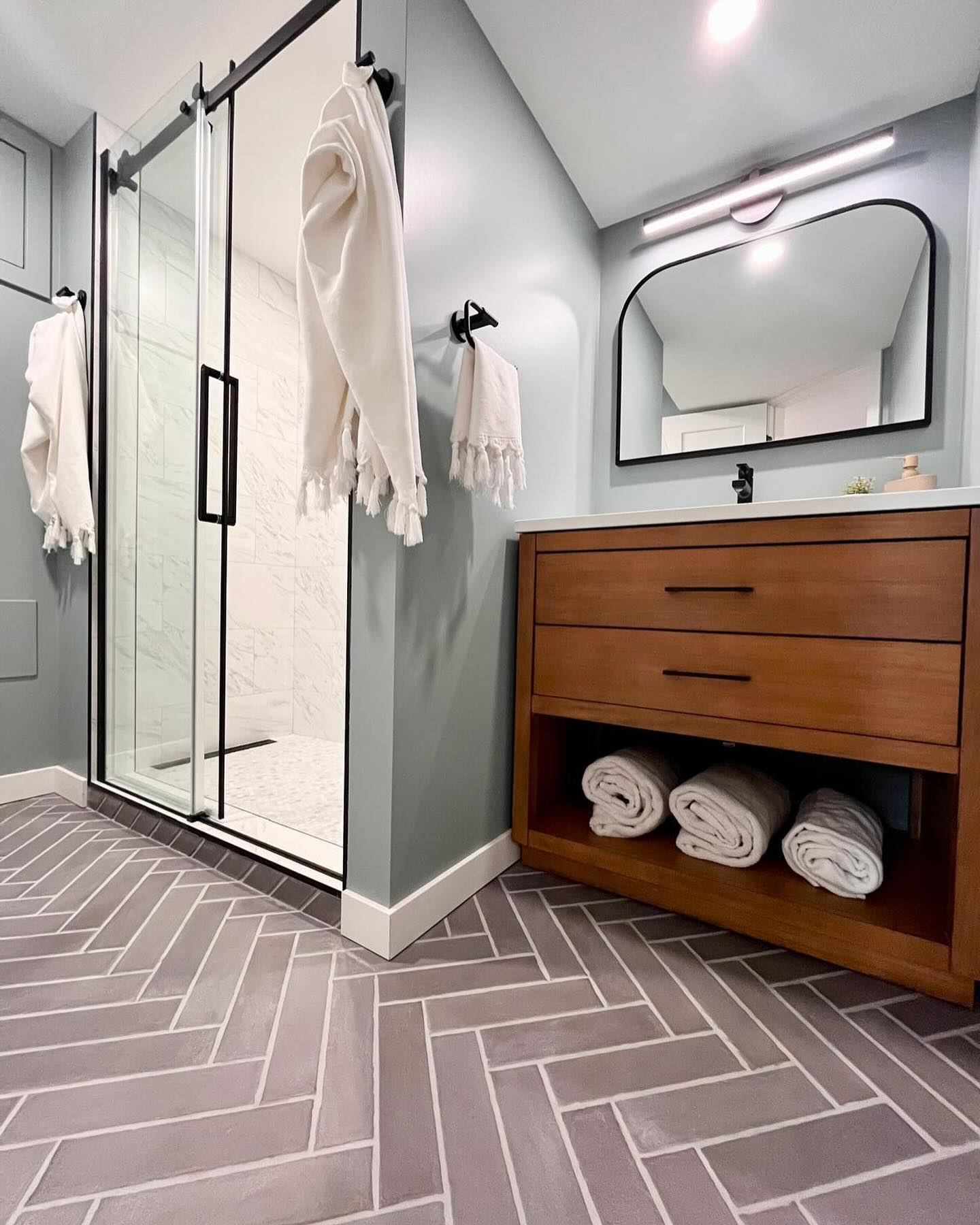 Bathroom with herringbone floor tile, a wood vanity, and a black-framed glass shower