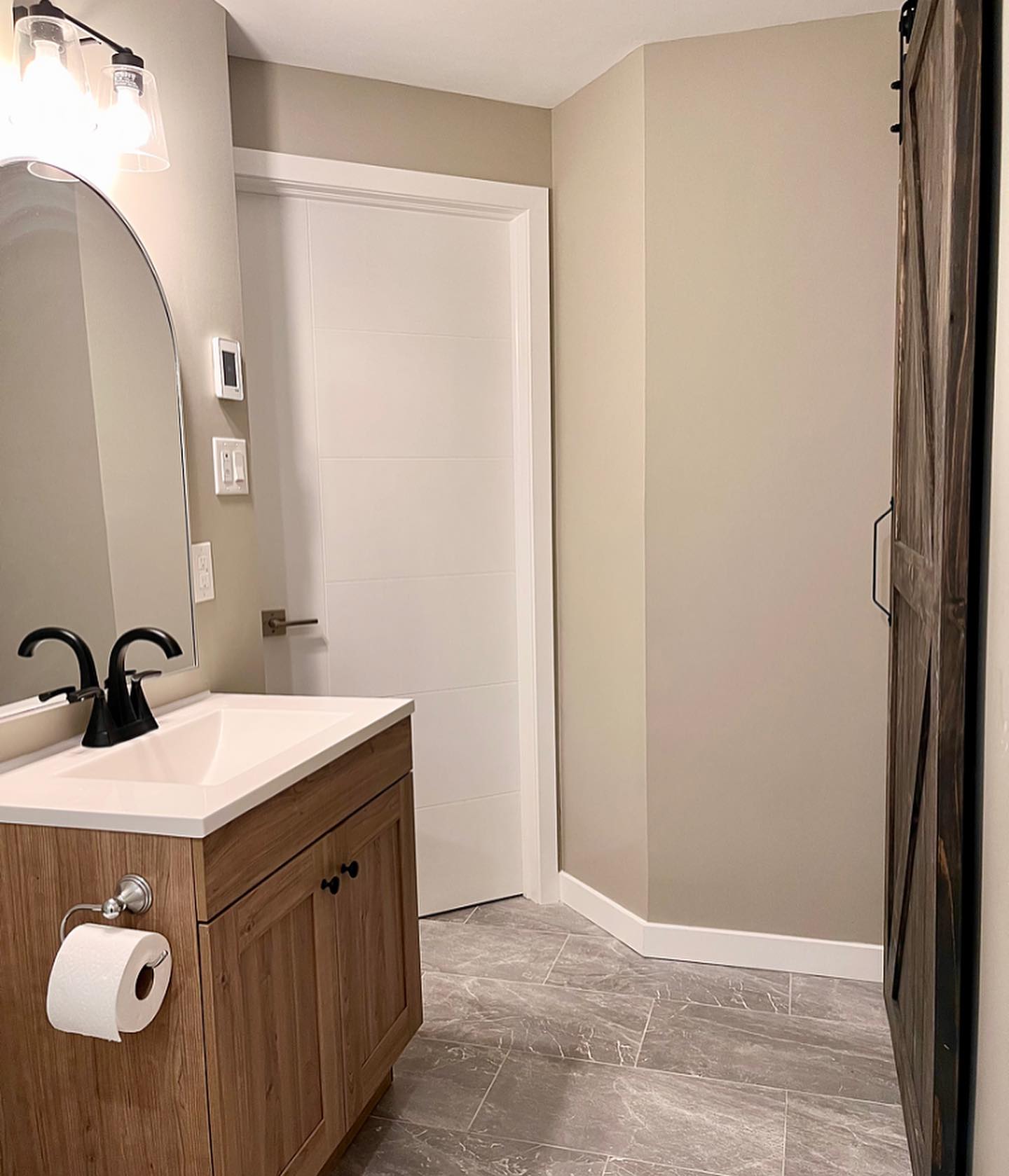 Bathroom vanity in light wood with a white sink, arched mirror, and a dark barn door